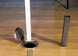 Badminton net post in floor sleeve with open lid, next to a square metal post on a wooden floor.