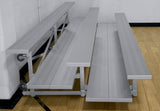 Two-row aluminum bleacher with double foot plank and wheels, set against a white brick wall and wood floor.