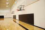 Black wall pads installed in a gymnasium with basketball hoops and a wooden floor.