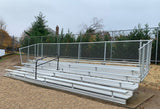 Aluminum bleacher with 5 rows, a black handrail, and chain-link fencing, on a dirt and grass field.