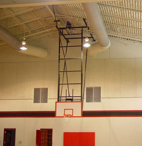 Ceiling-mounted basketball backstop with a white backboard and orange rim in a gymnasium.