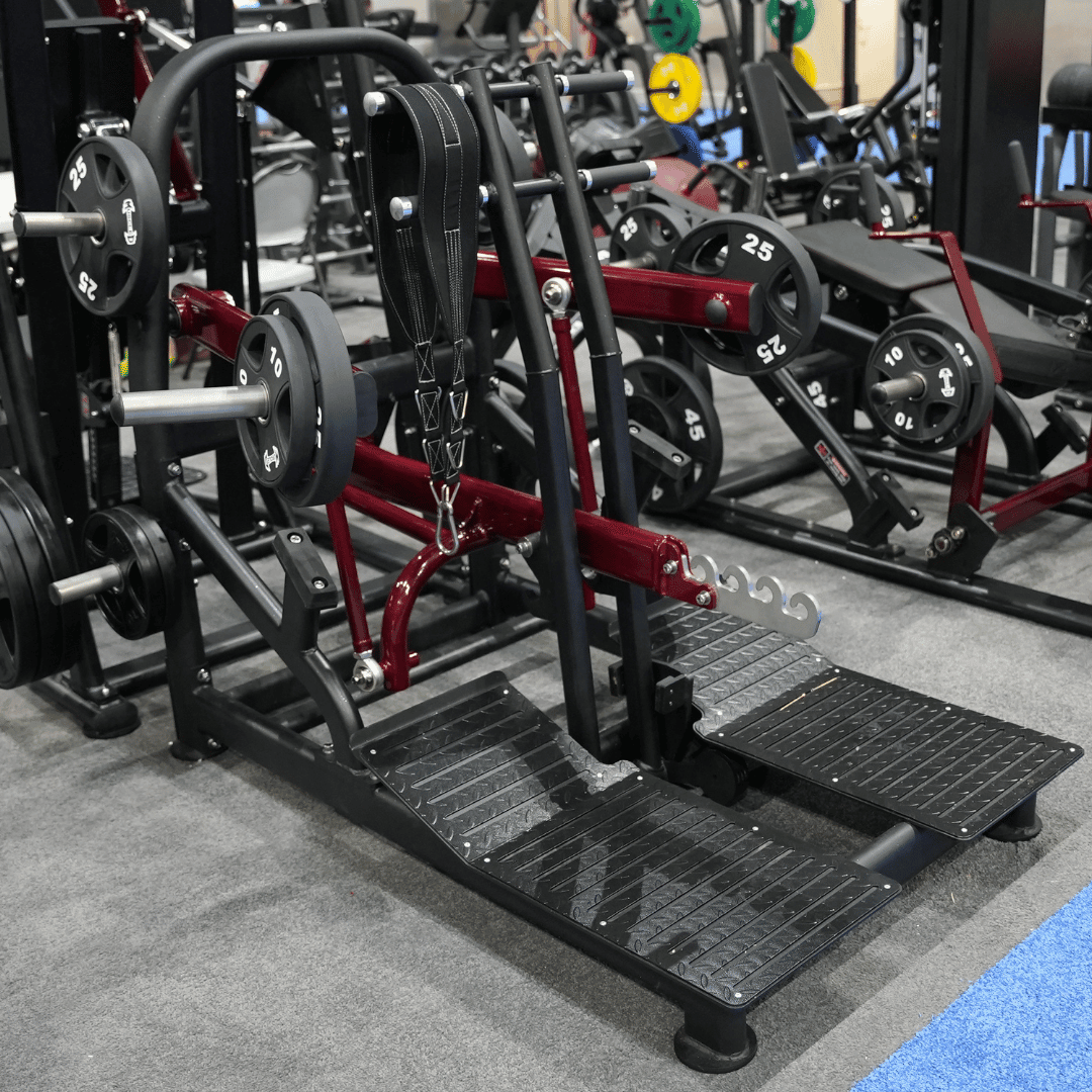 Plate-loaded belt squat machine with a black belt and foot platforms, in a gym setting.