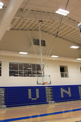 Ceiling-hung basketball backstop with a clear backboard and orange rim, in a gym with blue bleachers.