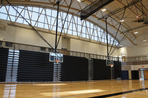Ceiling-hung basketball backstop with a clear backboard and orange rim in a gym with bleachers.