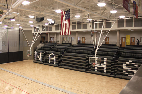 Two ceiling-hung basketball backstops with bent single posts in a gymnasium with bleachers.