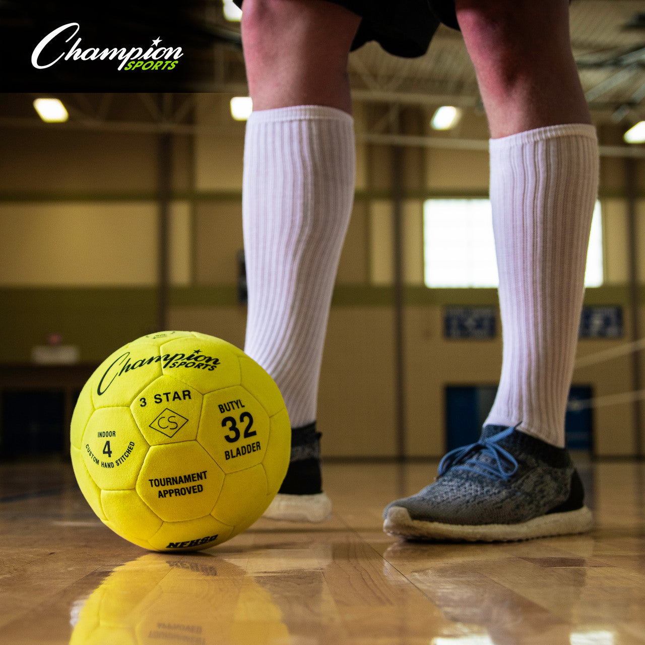 Yellow Champion Sports 3-Star indoor soccer ball on a gym floor with a player's legs in the background.