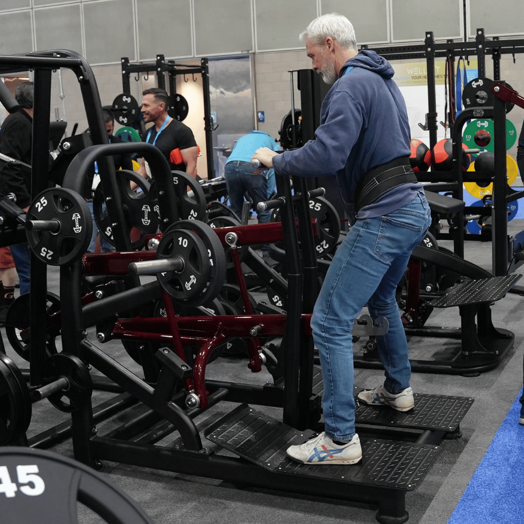 Man in jeans and a belt squat machine with weight plates in a gym setting.