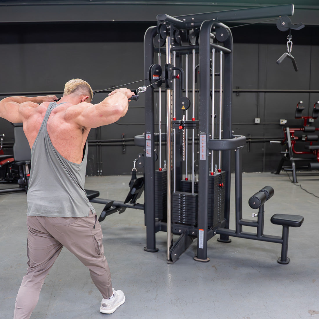 Man in gray tank top using a 4-stack jungle gym with cable pulleys for a rear delt exercise.