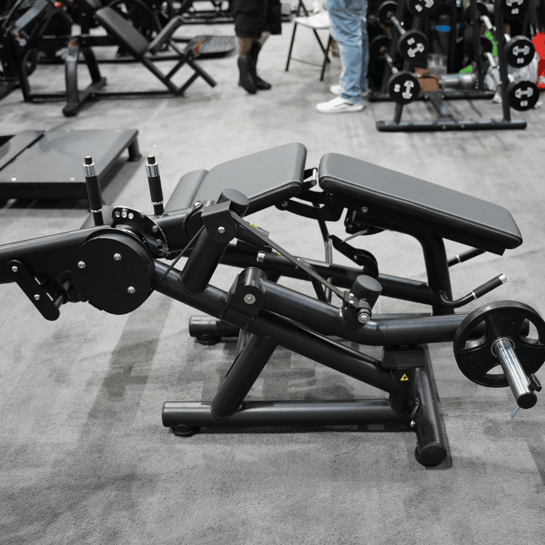 Black plate-loaded leg extension/curl machine with gray pads in a gym setting.