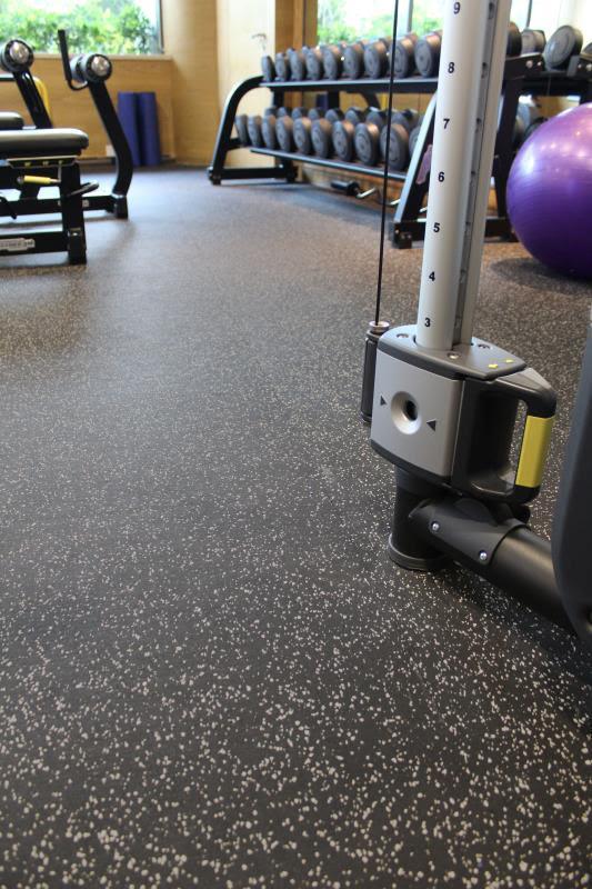 Black gym flooring with gray flecks in a gym setting with weights and exercise equipment.