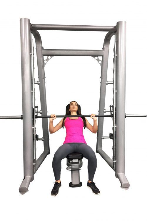Woman in pink tank top and gray leggings using a silver Smith machine with a black barbell.