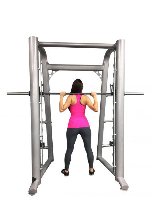 Woman in pink top and black leggings using a silver Smith machine with a barbell on her shoulders.
