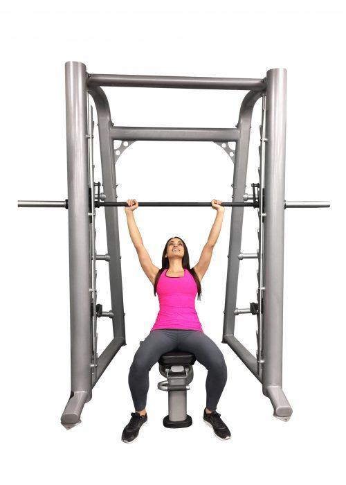Woman in pink top and gray leggings using a silver Smith machine with a black barbell.