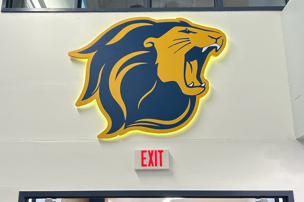 Backlit acrylic sign of a navy and gold roaring lion head on a white wall above an exit sign