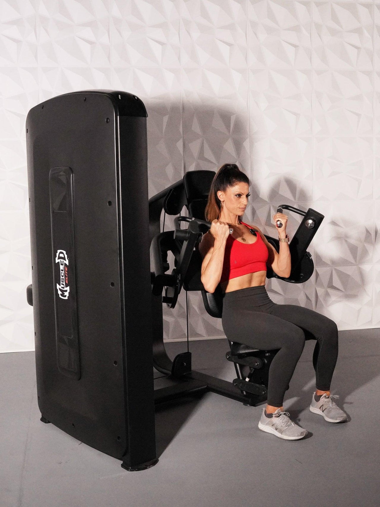 Woman in red top and black leggings using a black biceps curl machine with a white textured wall background.