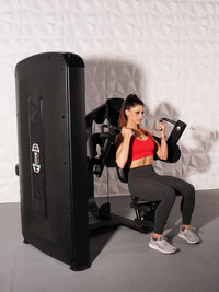 Thumbnail for Woman in red top and black leggings using a black biceps curl machine with a white textured wall background.