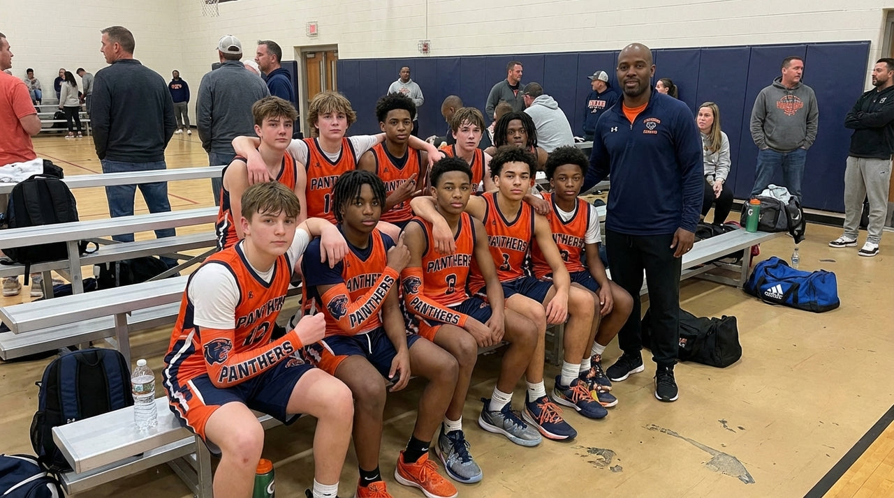 Basketball team in orange and navy "Panthers" jerseys, with a coach, sitting on benches in a gym.