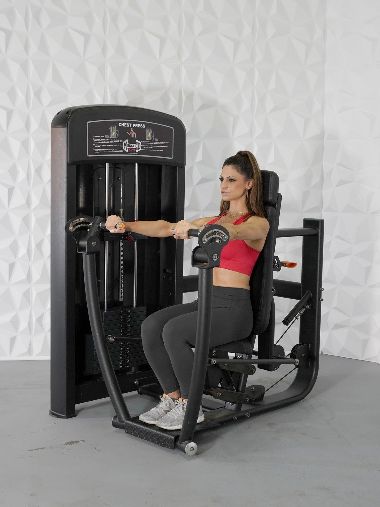 Woman in red top and black leggings using a black chest press machine with a weight stack.