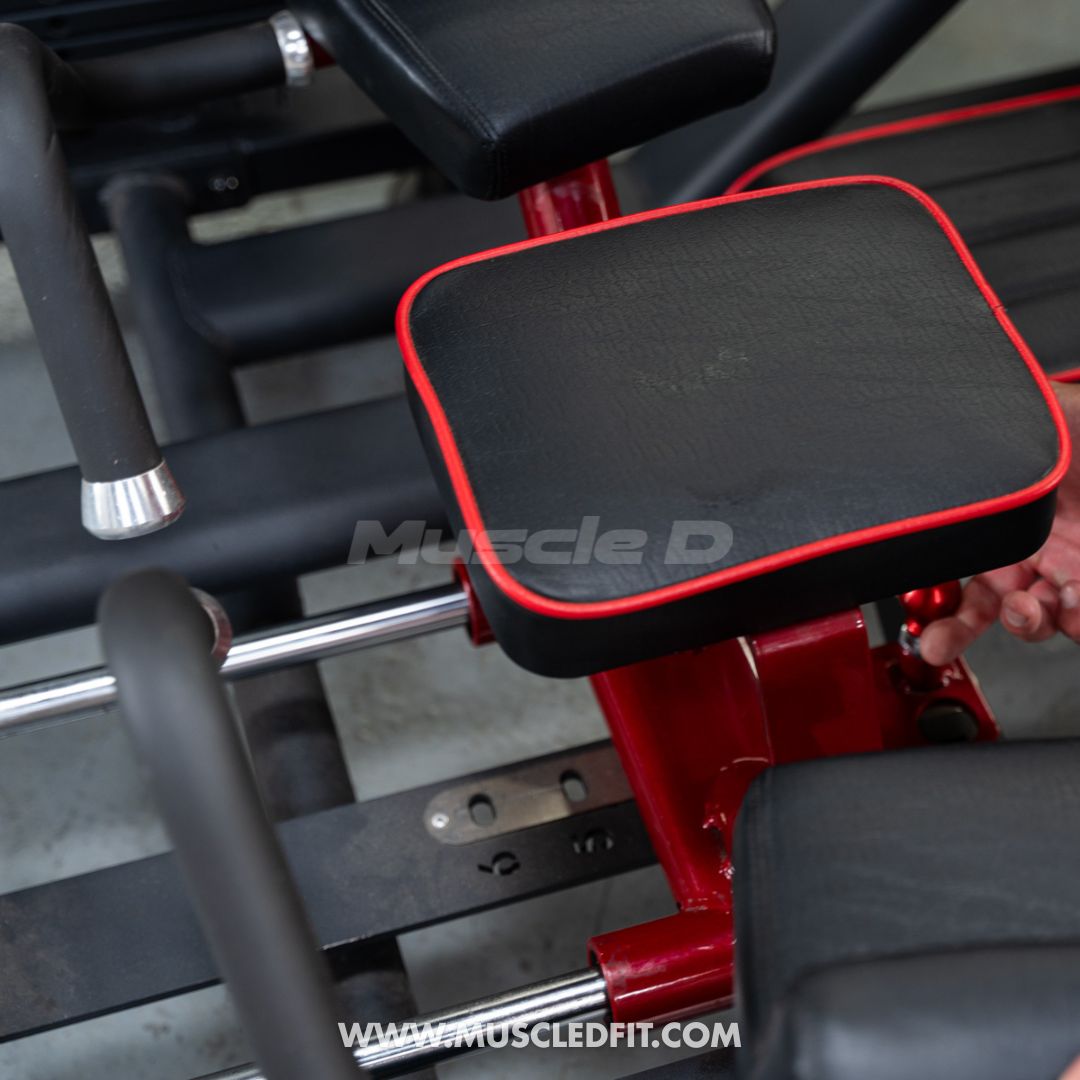 Close-up of a glute kick machine with black pads and red trim, showing part of the frame and a hand adjusting it.