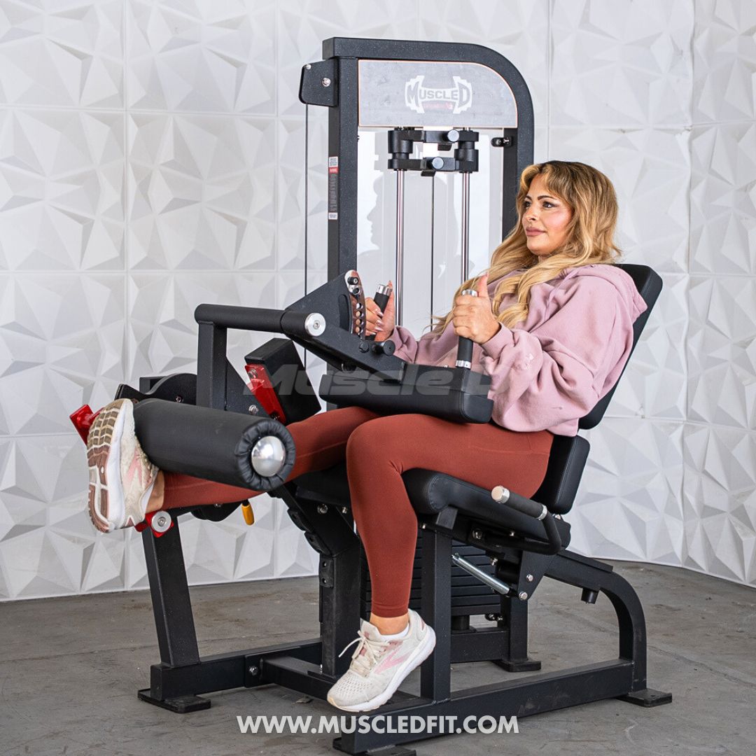 Woman using a black selectorized leg extension/seated leg curl combo machine against a white textured wall.