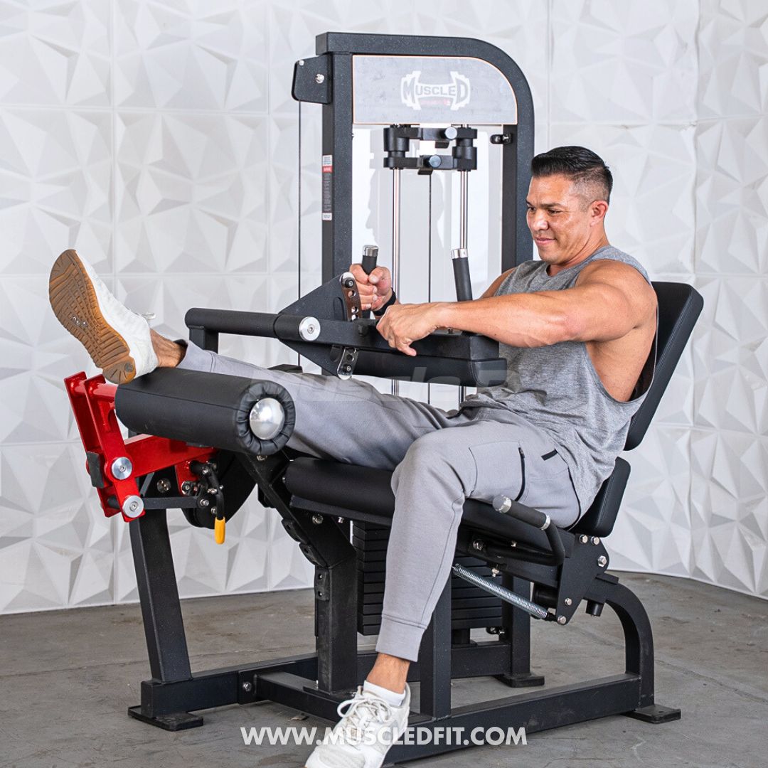Man in gray tank top and sweatpants using a black leg extension/curl machine with red accents.