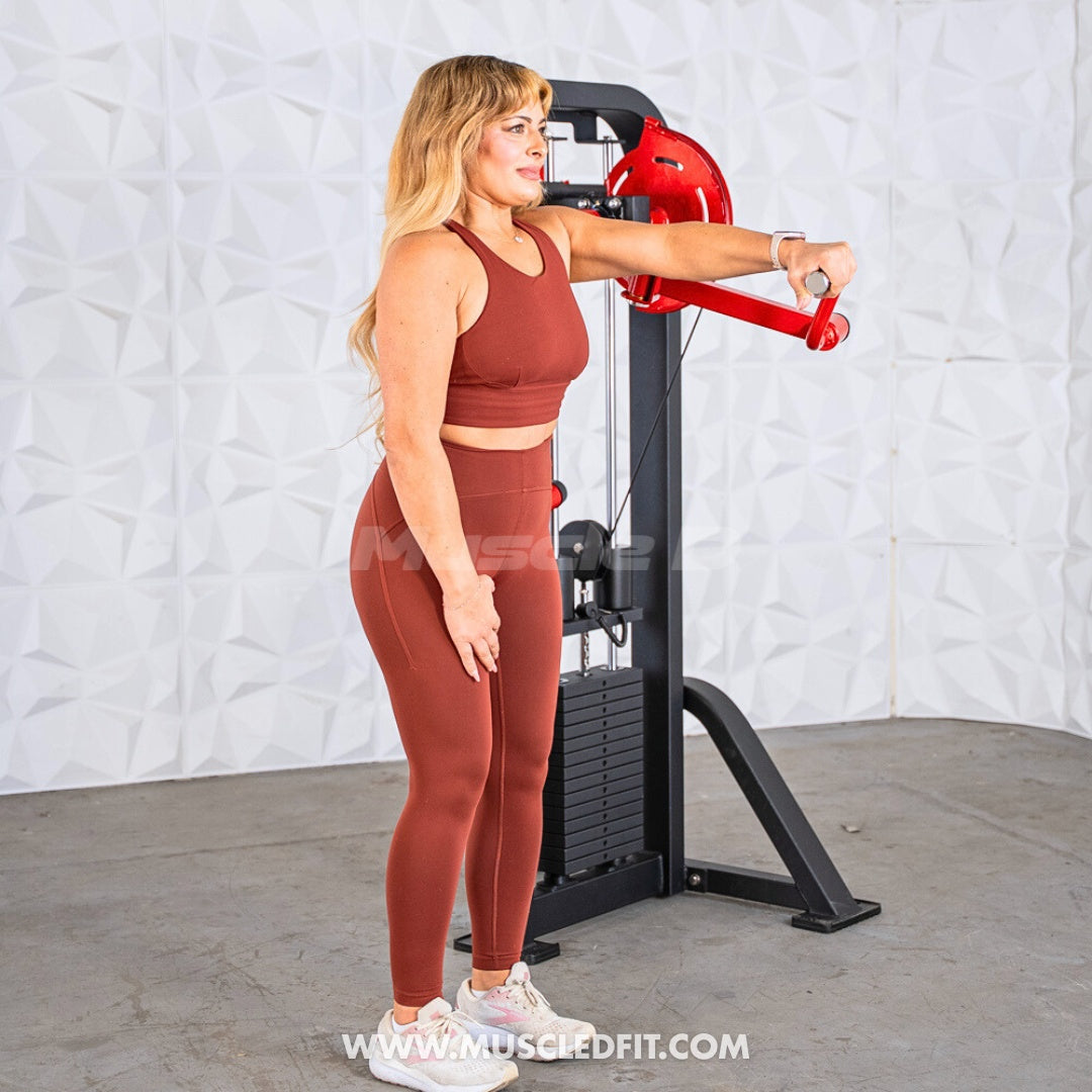Woman in rust-colored workout clothes using a black and red lateral raise machine.