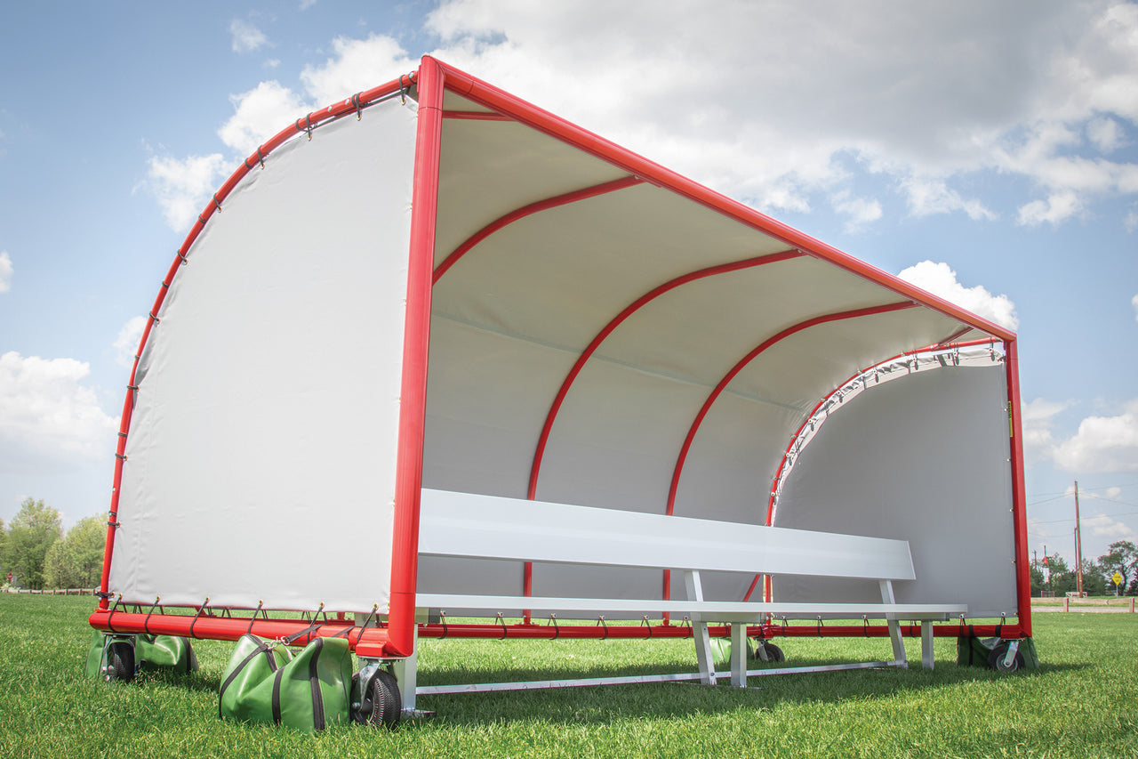 Portable sports team shelter with a white cover, red frame, bench and green sandbags on a grass field