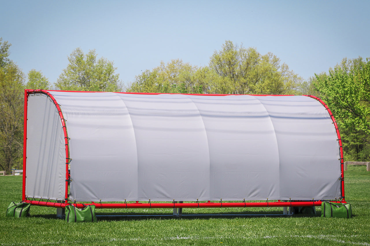 Rear view of a portable team shelter with a white cover and red frame weighted by green sandbags on grass