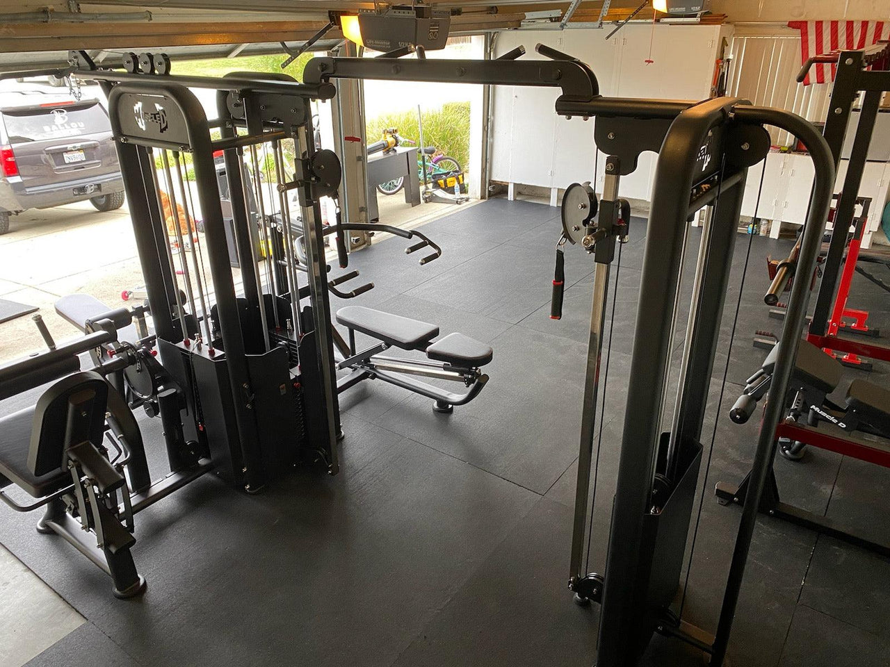 Black 5-stack multi-gym with benches and various attachments on a black gym floor in a garage.