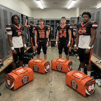 Thumbnail for Four football players stand in a locker room with their customized orange and black duffle bags.