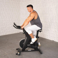 Thumbnail for Man in gray tank top and white pants riding a black upright exercise bike.