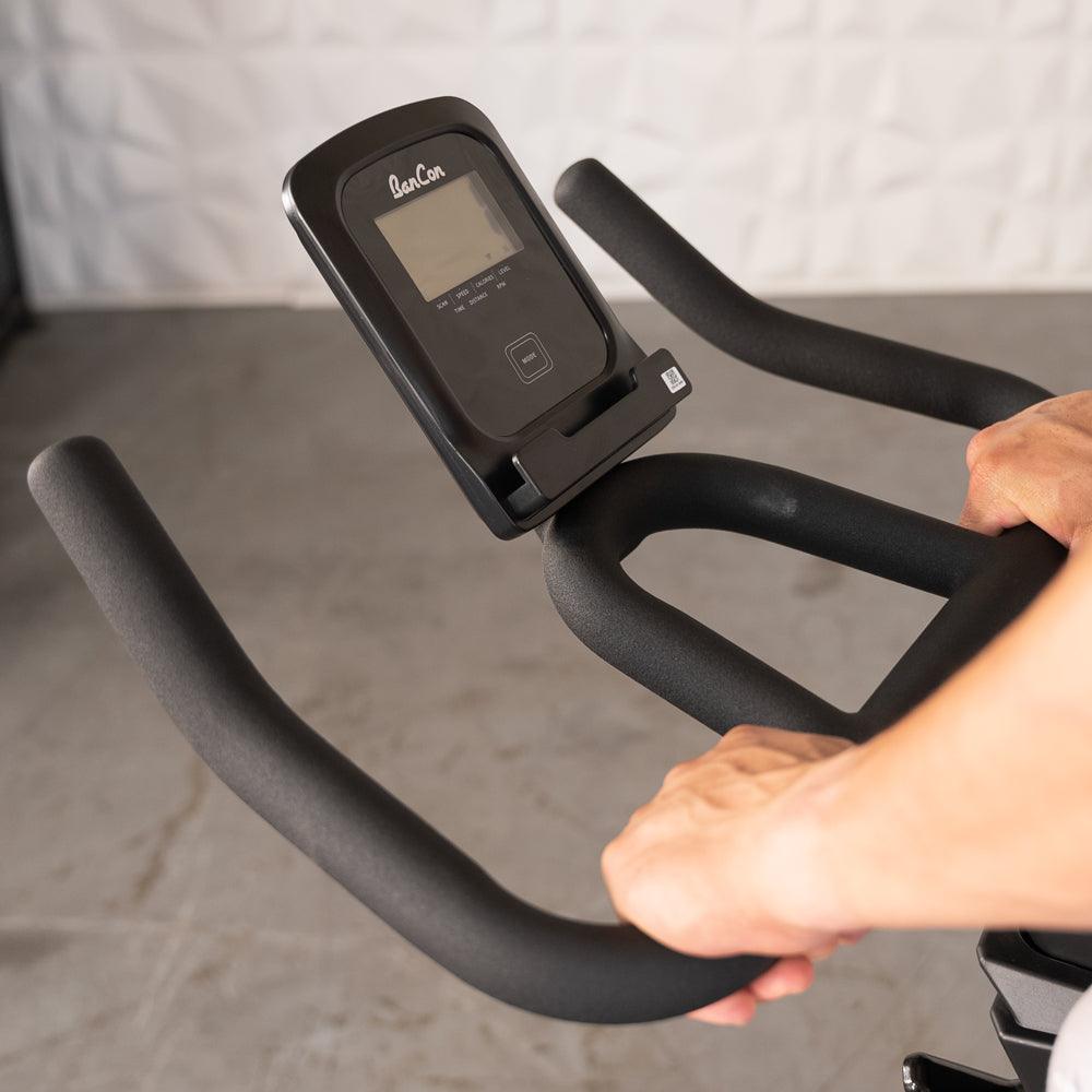 Person's hands gripping the black foam handlebars of an exercise bike with a digital display.