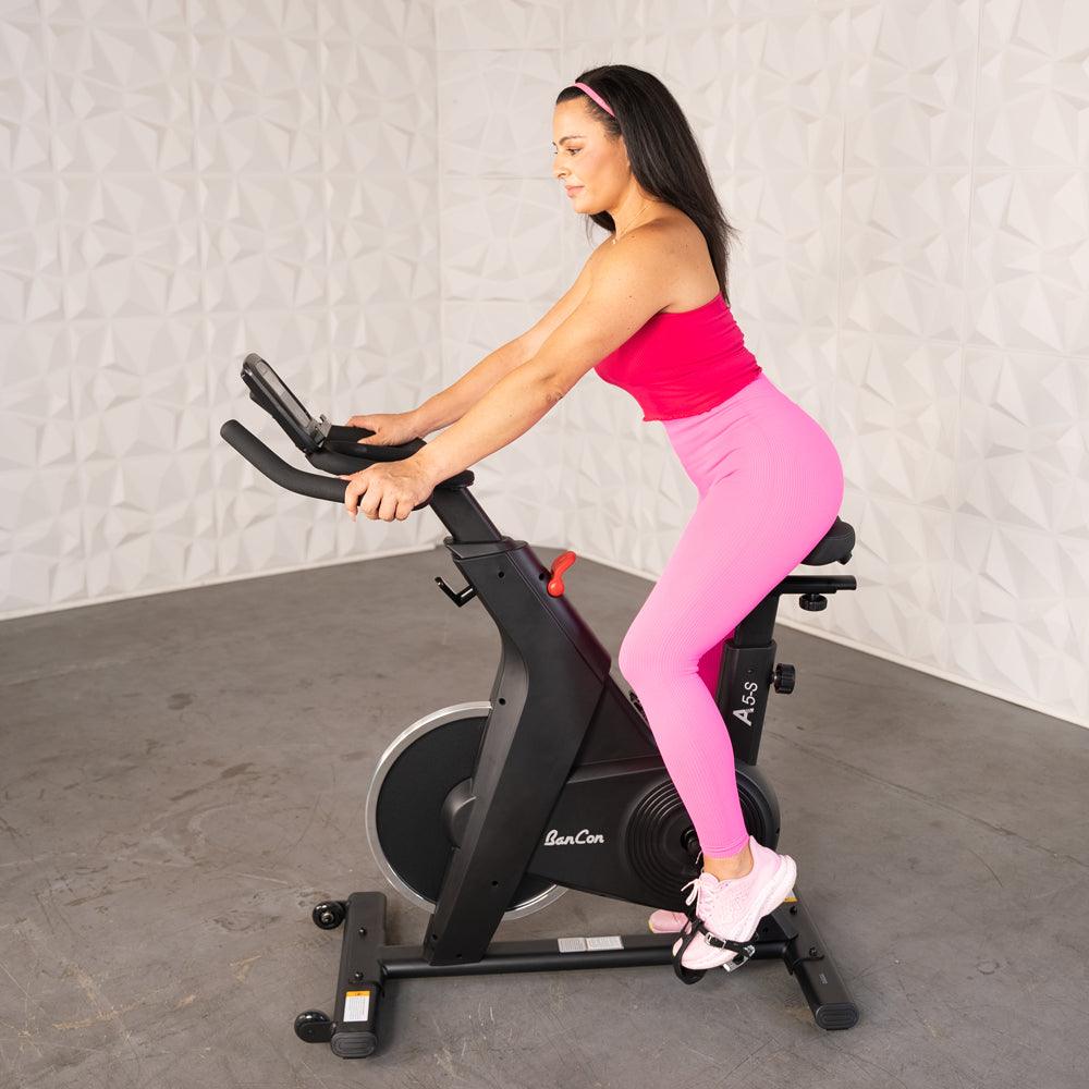 Woman in pink activewear riding a black upright exercise bike with a screen and adjustable features.