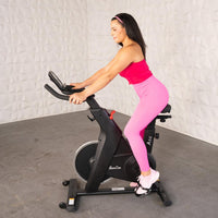 Thumbnail for Woman in pink activewear riding a black upright exercise bike with a screen and adjustable features.