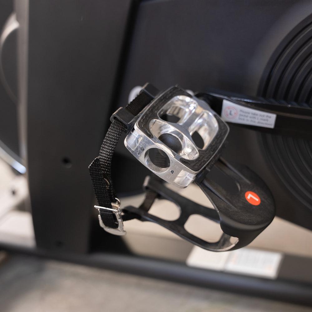 Close-up of an exercise bike pedal with a clear toe cage and black strap.