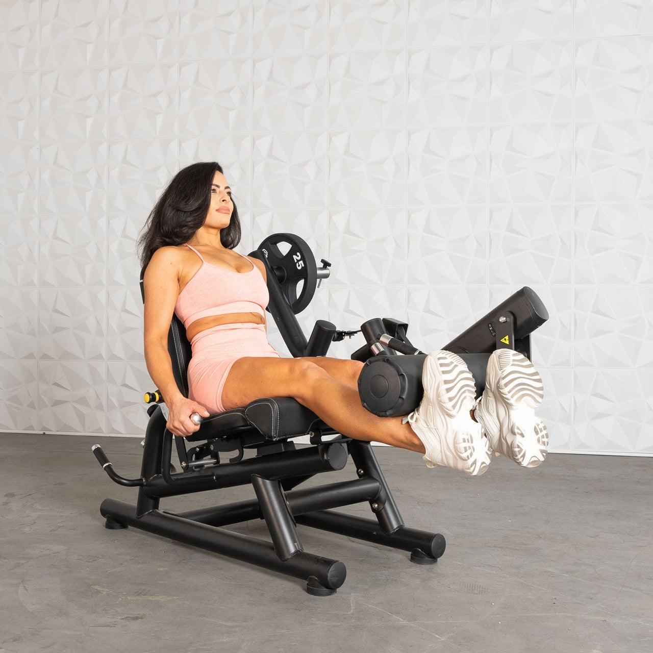 Woman in pink activewear using a black leg extension/curl machine with a 25lb plate.