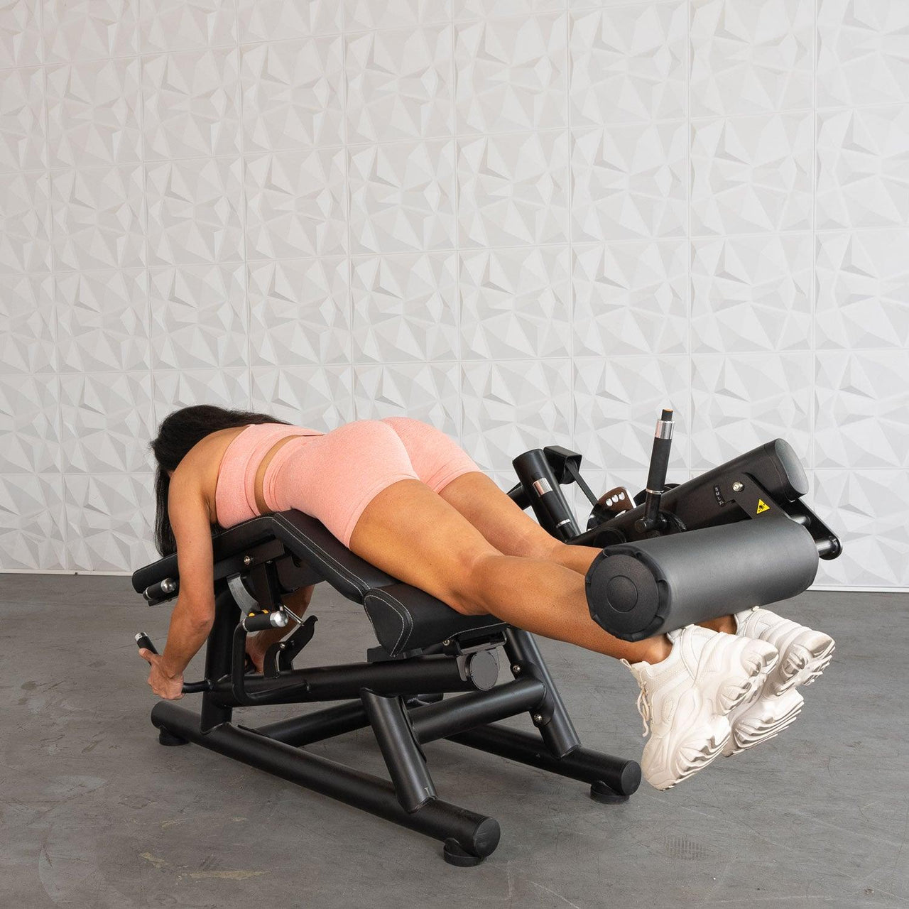 Woman in peach workout gear using a black leg curl machine, lying prone with feet under the roller.
