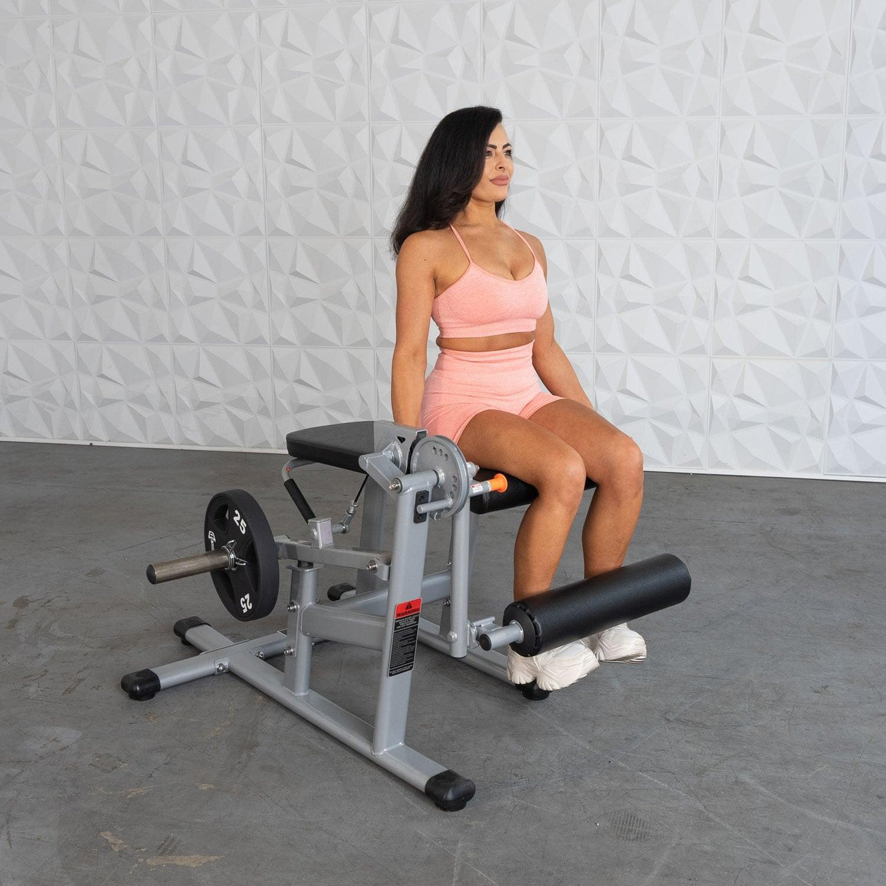 Woman in peach activewear sitting on a silver and black leg extension/curl machine with a weight plate.