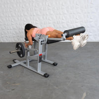 Thumbnail for Woman in pink doing leg curls on a silver and black plate-loaded fitness machine.