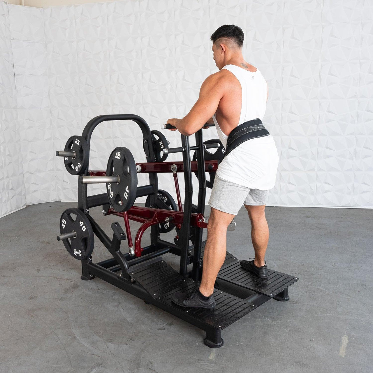 Man in a white tank top and grey shorts using a plate-loaded belt squat machine with 45lb weights.