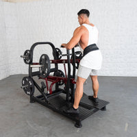 Thumbnail for Man in a white tank top and grey shorts using a plate-loaded belt squat machine with 45lb weights.
