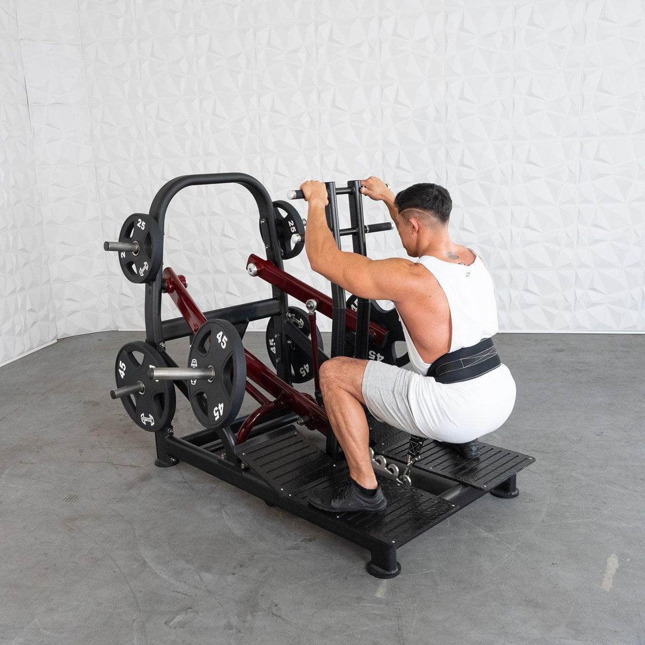 Man in white tank top and shorts performing a belt squat on a plate-loaded machine.