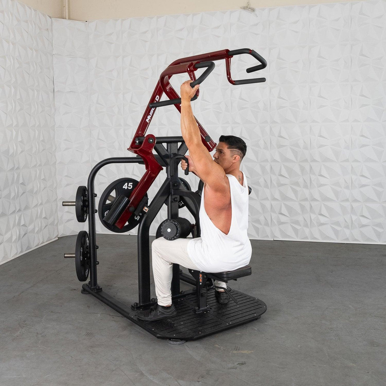Man in white tank top using a red and black rotary lat pull-down machine with 45lb plates.