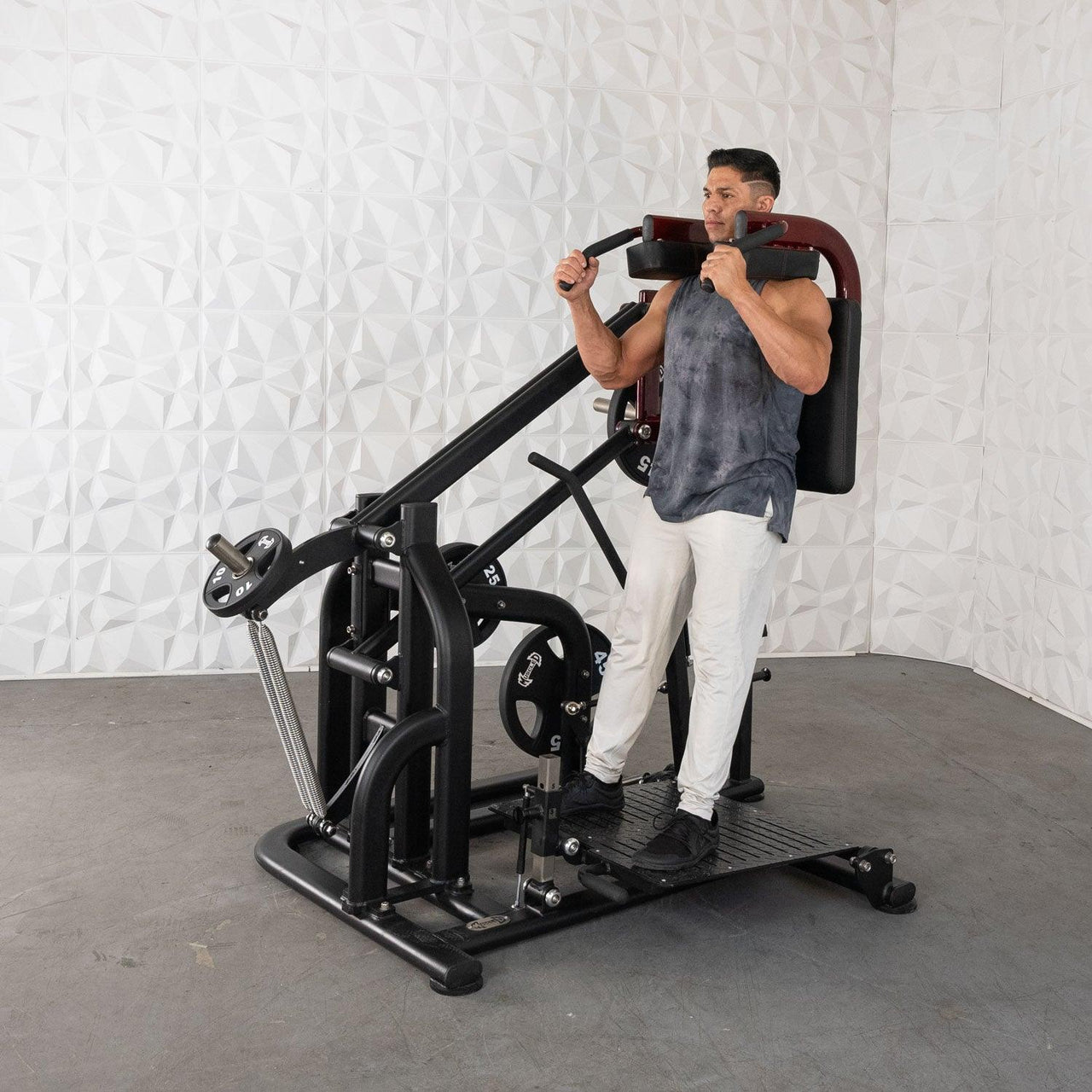 Man using a black plate-loaded squat machine with red shoulder pads and a textured white wall in the background.