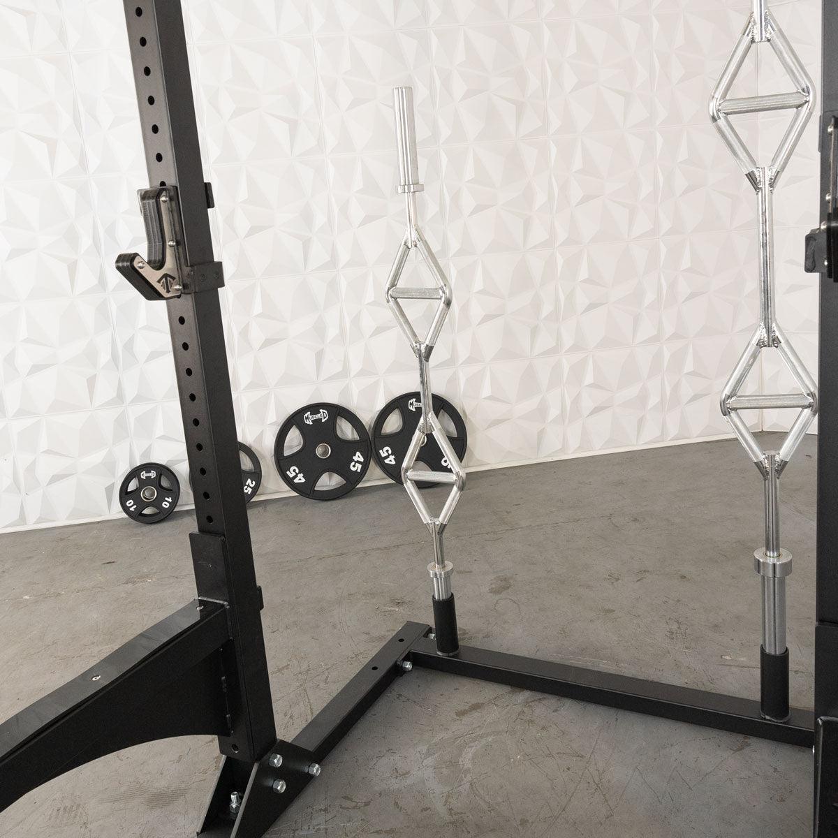 Black squat stand with J-hook, two chrome trap bars, and several weight plates on a concrete floor.