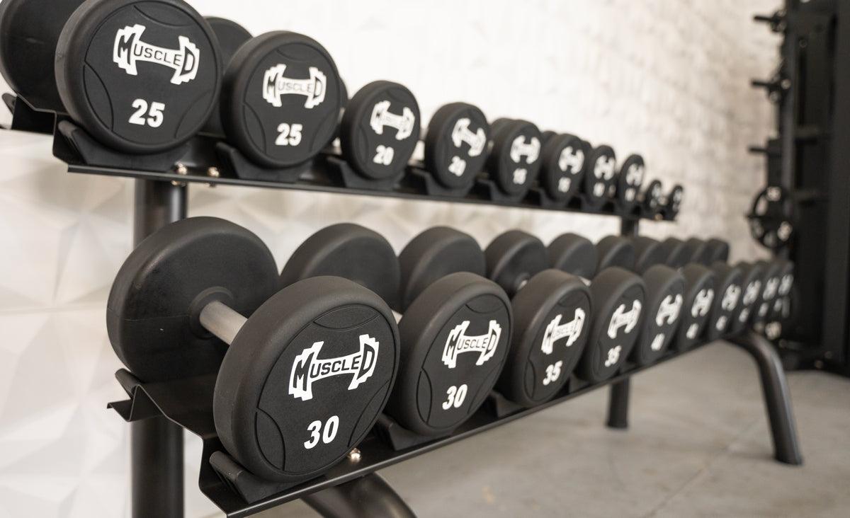 Black urethane dumbbells with "Muscle D" logo and weight numbers on a two-tier rack in a gym.