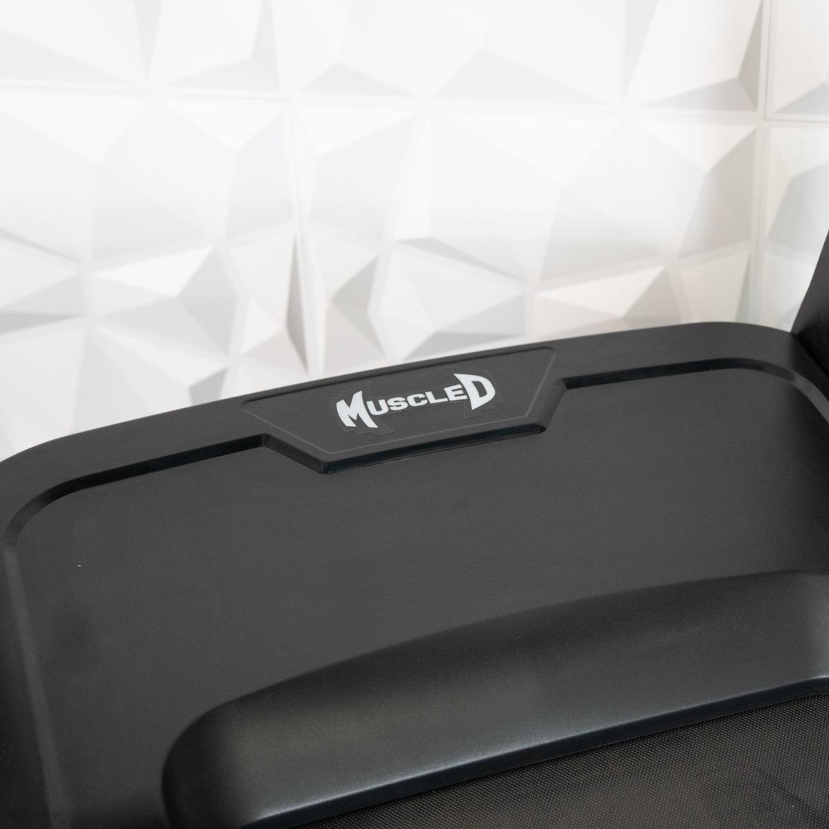 Close-up of a black treadmill's console with "Muscle D" logo in white, against a white geometric wall.