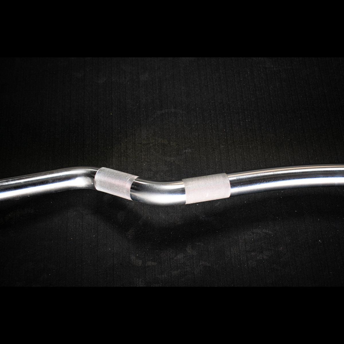 Close-up of a silver, hard chrome curl bar with textured grip sections against a dark background.