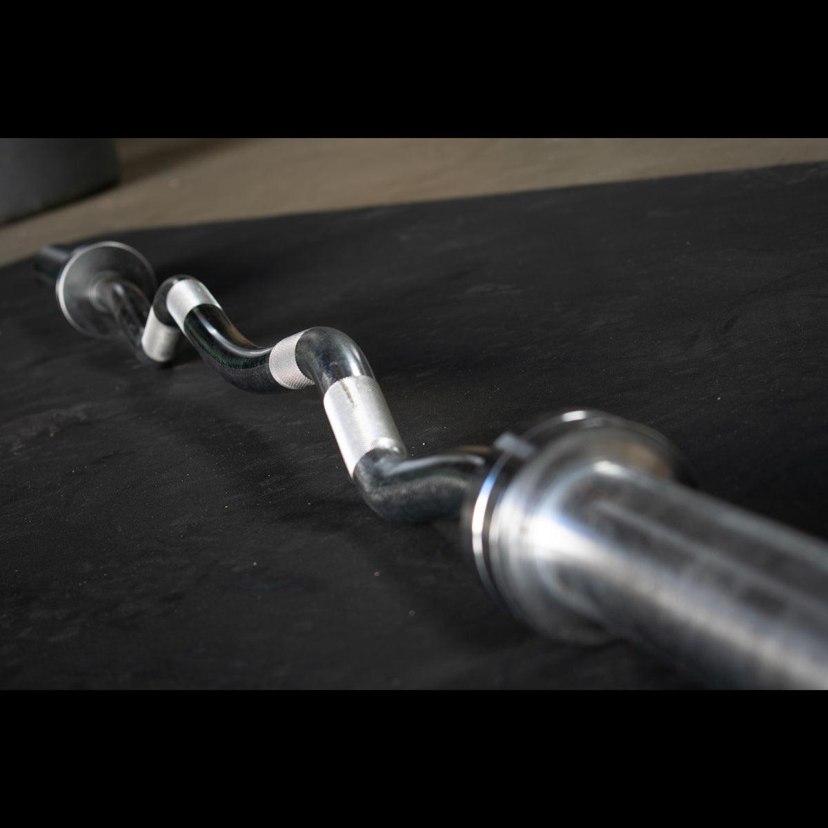 Hard chrome curl bar with ergonomic angles on a dark surface.