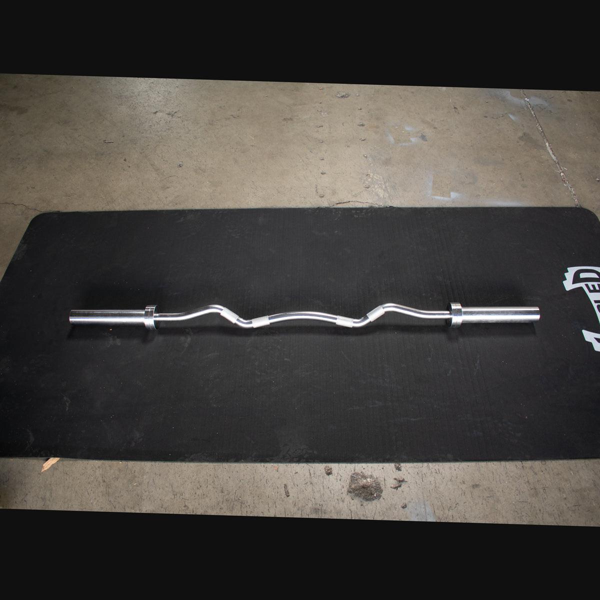 Silver hard chrome curl bar with ergonomic angles, lying on a black mat.
